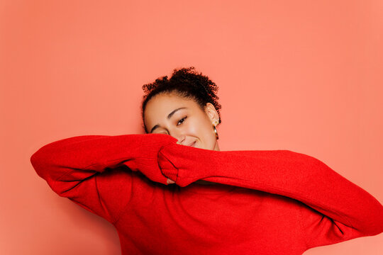Smiling woman covering face with red sweater against colored wall