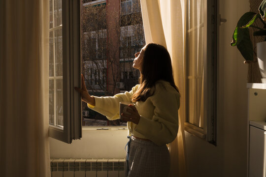 Woman enjoys morning light while standing by window with coffee