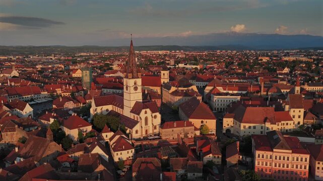 Aerial 4k video of the old town of Sibiu featuring historic red roofs and the tall church tower, with the majestic blue Fagaras mountains visible on the horizon. Sibiu, Transylvania, sunset landscape.