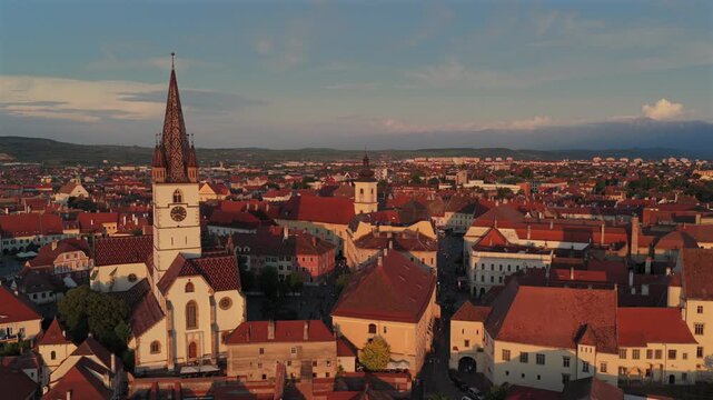 Aerial 4k video of the old town of Sibiu featuring historic red roofs and the tall church tower, with the majestic blue Fagaras mountains visible on the horizon. Sibiu, Transylvania, sunset landscape.