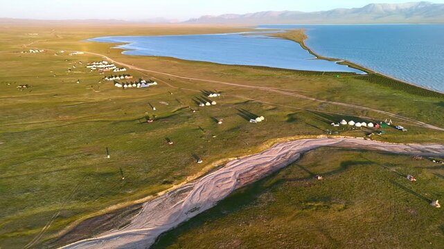 Aerial view of Song-Kul lake with traditional yurt camps scattered across the green plains under golden hour light in Naryn Region, Kyrgyzstan.