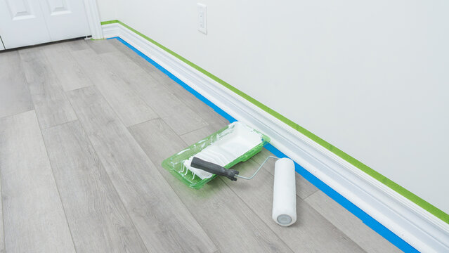 Home renovation scene featuring paint roller, tray, and painter's tape on freshly prepared floor with white walls and door frame visible