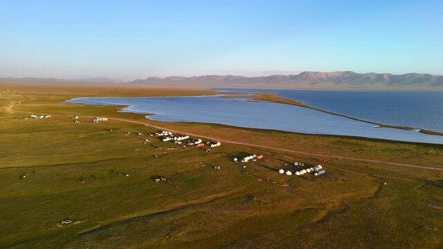 Aerial view of Song-Kul lake with traditional yurt camps scattered across the green alpine pastures under a clear sky in Naryn Region, Kyrgyzstan.