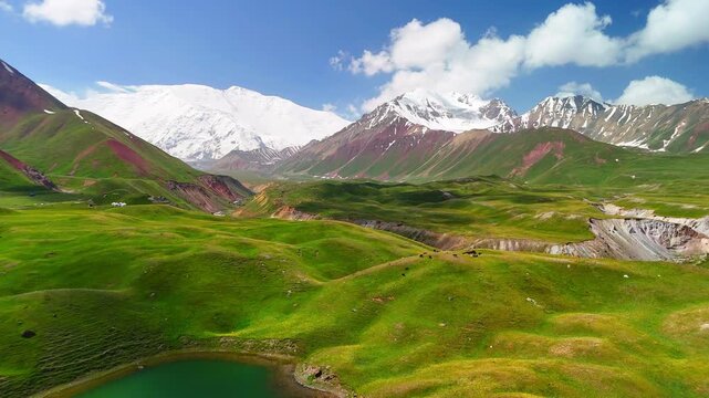 Aerial view of Tulpar Lake surrounded by green hills and snow-capped Pamir Mountains in Alay District, Osh Region, Kyrgyzstan.