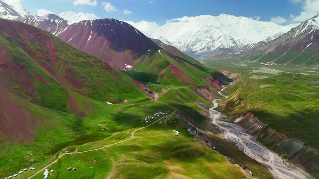 Aerial view of yurt camps nestled in the green Alay Valley with a winding river and snow-capped Pamir Mountains in Alay District, Osh Region, Kyrgyzstan.