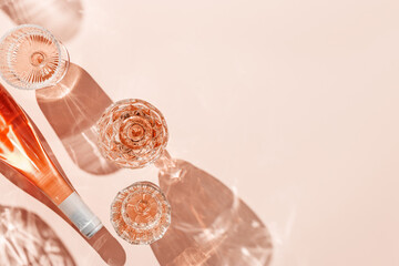 Fototapeta premium Wine Glasses and bottle of rose wine on pink background, sun glare and reflection from sun. Aesthetic composition from crystal glasses, sparkling alcohol drink, champagne, Summer romantic setting