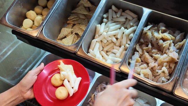 Close-up of hands using tongs to select various hot pot ingredients from stainless steel trays