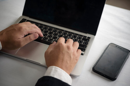 Businessman using laptop for data entry and communication with smartphone on desk. Focused on fintech, cybersecurity, and modern workspace efficiency in close-up shot.