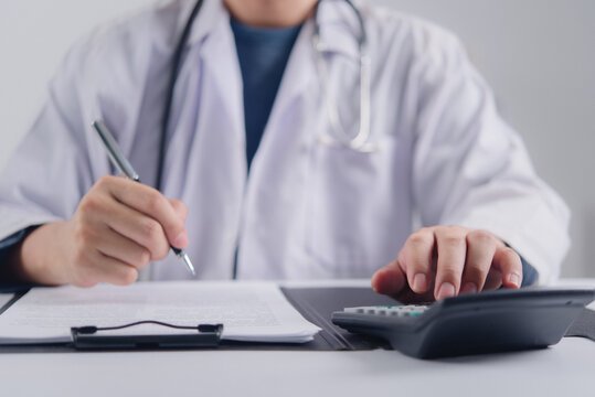 Professional physician in white coat calculating medical fee evaluating health insurance premium and writing clinical report on paperwork showing healthcare financial management concept