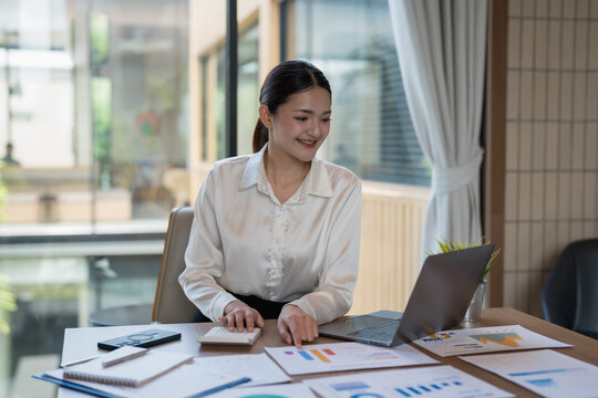 Asian businesswoman analyzing financial charts and data on papers and laptop while calculating figures with a calculator, working proactively in a modern office setup