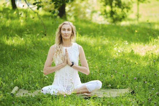 A woman dressed in white sits cross-legged on a mat, practicing yoga outdoors. Surrounded by vibrant green grass and trees, she has her hands in a prayer position, embodying peace and tranquility