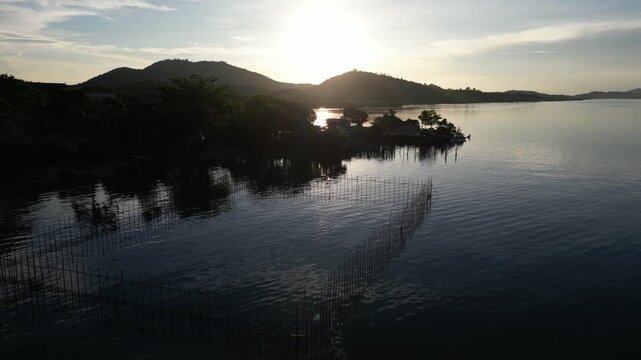 Aerial view of a traditional fish trap in the calm waters near lush hills during a golden sunset in Riau Islands, Indonesia.