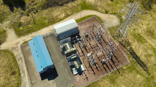 High angle drone footage showing an electrical power substation with transformers and high voltage transmission towers, surrounded by a rural landscape on a sunny day with distinct shadows