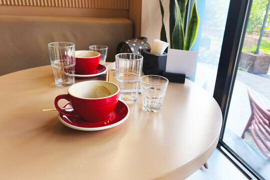Dirty empty red coffee cup and clear glasses left on a cafe table after visitors in soft daylight with a cozy casual atmosphere