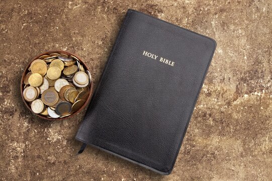 Holy Bible and bowl of coins on a textured stone surface