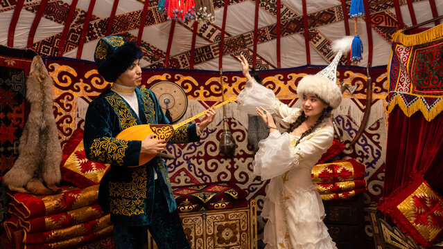 Kazakh man playing dombra while woman in traditional national clothing dances inside a yurt, showcasing authentic cultural heritage of Kazakhstan.