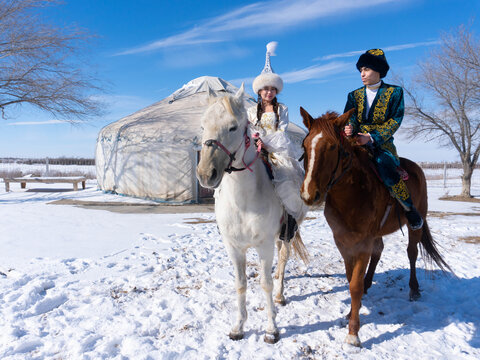 Kazakh man and woman in traditional clothing standing with horses near a yurt, representing nomadic culture and heritage of Kazakhstan.