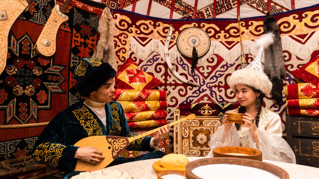 Kazakh couple in traditional clothing enjoying kumis while man plays dombra inside a yurt, showcasing authentic nomadic culture.