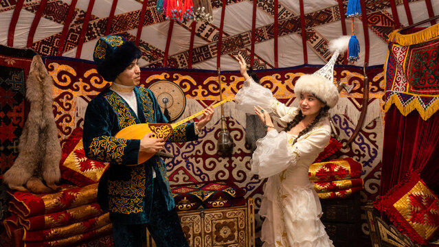 Kazakh man playing dombra while woman in traditional national clothing dances inside a yurt, showcasing authentic cultural heritage of Kazakhstan.