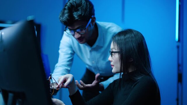 Tech engineers collaborate on a modern robotics project in a high-tech lab at night, analyzing code on multiple monitors and testing a prototype robotic arm component under ambient blue neon lights.