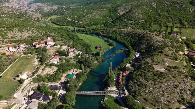 Aerial view of the turquoise Zrmanja river winding through a lush green valley with a small bridge and traditional houses in Muskovci, Zadarska zupanija, Croatia.