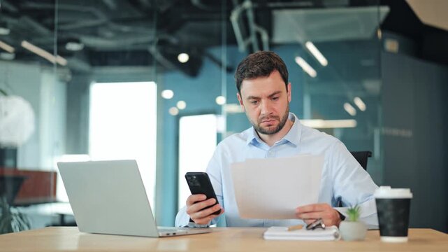 Focused businessman or IT specialist checking corporate documents and typing on smartphone. Happy corporate worker, manager or freelance designer smiling at camera at contemporary coworking desk.