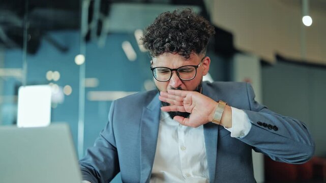 Exhausted male professional in business suit glasses sitting at workplace yawning stretching arms. Corporate burnout stress exhaustion ending workday concept. Professional worker needing rest.