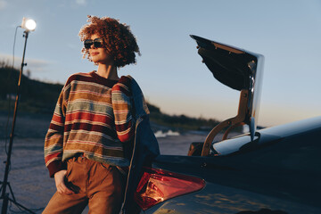 Fototapeta premium Woman lifestyle portrait with curly hair wearing sunglasses beside a car trunk, laptop open with a graduation cap, outdoors at sunset, casual travel mood, soft evening light, natural breeze