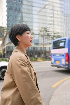 Young woman walks city street, looking ahead. Urban buildings loom behind her, blurred in distance. She wears a beige coat, short dark hair. Blue bus passes by, adding motion to the scene