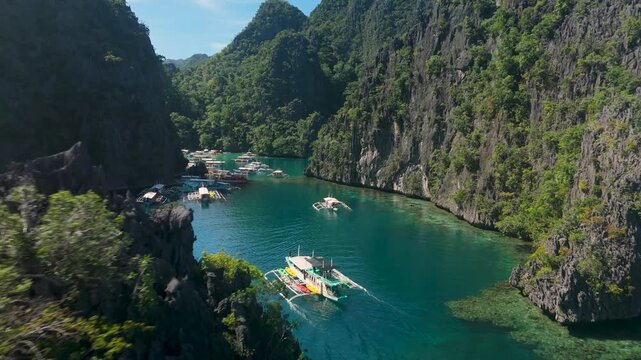 Aerial flypass beside bangka outrigger tour boats arriving at turquoise Kayangan Lake entrance surrounded by limestone karst cliffs in tropical paradise of Coron Palawan Philippines