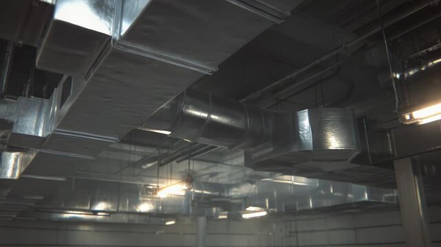 Overhead view of an industrial ceiling showcasing a complex network of galvanized metal ventilation ducts, pipes, and fluorescent lighting in a warehouse or factory setting