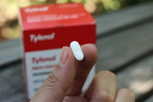 Hand holding white caplet medicine pill with a blurred pharmaceutical box in the background