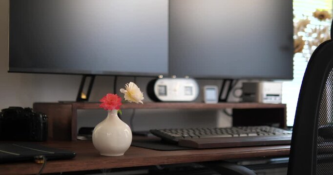 Cinematic vertical close up of a white ceramic flower vase with fresh roses on a wooden computer desk, blurred dual monitor setup background