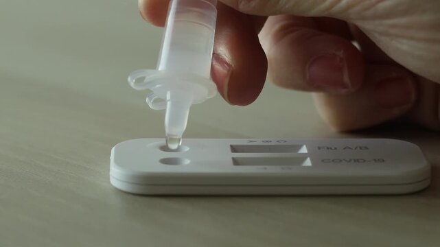 Close-up of a hand dropping liquid onto a rapid antigen test kit for Covid-19 and Flu A and B on a table, illustrating self-testing, medical screening, and healthcare diagnosis at home.