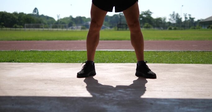 Cinematic medium close-up of a male athlete's lower body and legs doing a deep bodyweight squat on concrete at an outdoor stadium