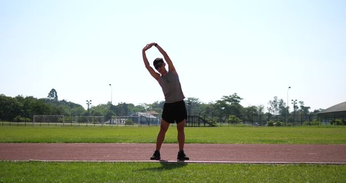 Cinematic front view wide shot of a male athlete warming up and doing a standing side bend stretch on an outdoor running track