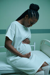 Fototapeta premium Vertical shot of pregnant Black woman sitting on hospital bed, holding belly and feeling discomfort during labor assessment. Ideal for maternity care, prenatal health, obstetrics marketing