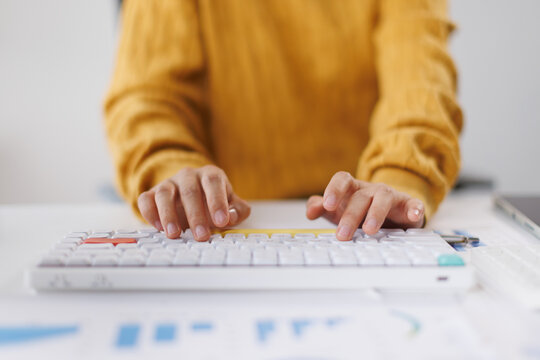 Person's hands typing on a modern white mechanical keyboard with colored keys, engaged in data entry, coding, or office work at a desk with blurred documents and a tablet