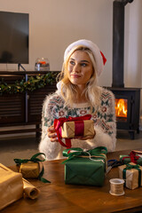 Obraz premium Woman wearing Santa hat and holiday sweater holding wrapped gift at wood table with scissors, stove
