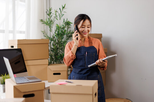 Female small business owner managing online orders, talking on phone and holding tablet, standing in home office fulfillment center surrounded by shipping boxes for e-commerce startup
