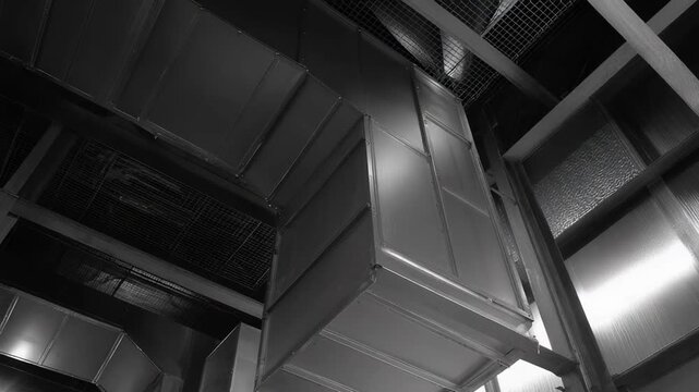 Intricate system of large metallic hvac ductwork and ventilation shafts spanning across the ceiling and walls of a modern industrial or commercial building interior, presented in black and white