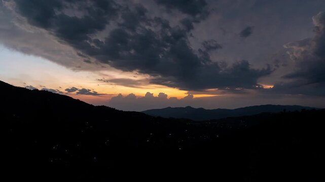 A high-altitude time-lapse capturing golden sunset light breaking through dramatic clouds over the silhouette of rolling mountain peaks in the serene Himachal Pradesh region.