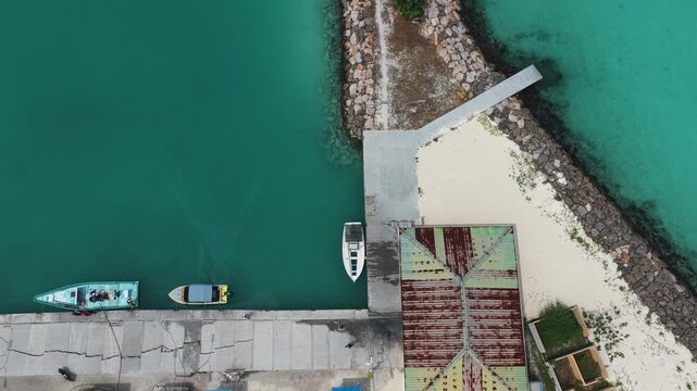 Aerial view of small boats moored at a concrete pier next to turquoise waters and a building with a rusty roof in Hanimaadhoo, Haa Dhaalu, Maldives.