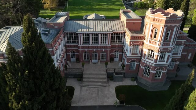 Aerial view of Chateau Mukhrani royal palace and estate in Georgia, magnificent pink castle architecture with a tower surrounded by a lush green park, gardens and pathways on a spring day.