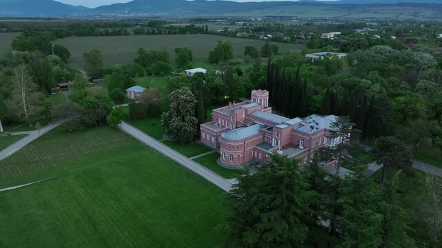 Aerial view of Chateau Mukhrani royal palace and estate in Georgia, magnificent pink castle architecture with a tower surrounded by a lush green park, gardens and pathways on a spring day.