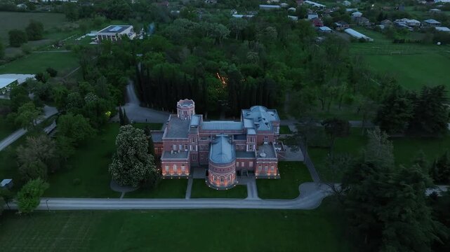 Aerial view of Chateau Mukhrani royal palace and estate in Georgia, magnificent pink castle architecture with a tower surrounded by a lush green park, gardens and pathways on a spring day.