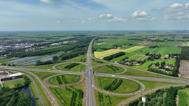 Aerial view of a highway cloverleaf interchange surrounded by green fields and industrial zones under a blue sky in Heerenveen, Friesland, Netherlands.