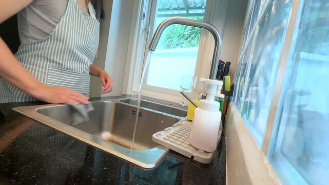 Woman wiping kitchen sink with cloth under running water. Real life video footage of home cleaning, washing sink, kitchen hygiene and everyday household chores.