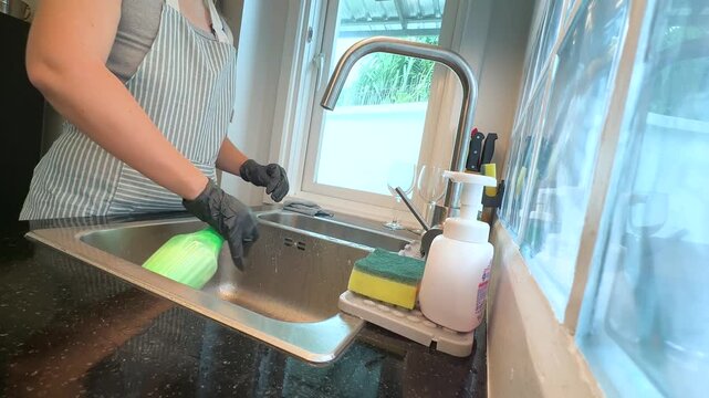 Woman spraying cleaning product and washing kitchen sink while wearing black rubber gloves. Real life video footage of home cleaning, kitchen hygiene, housework and everyday household routine.