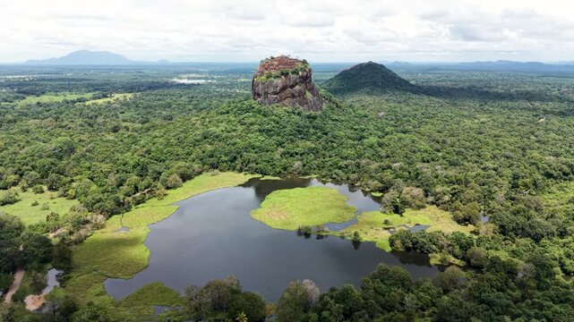 Aerial view of the ancient Sigiriya Rock fortress rising above the dense tropical jungle and a calm lake under a cloudy sky in Sigiriya, Central Province, Sri Lanka.